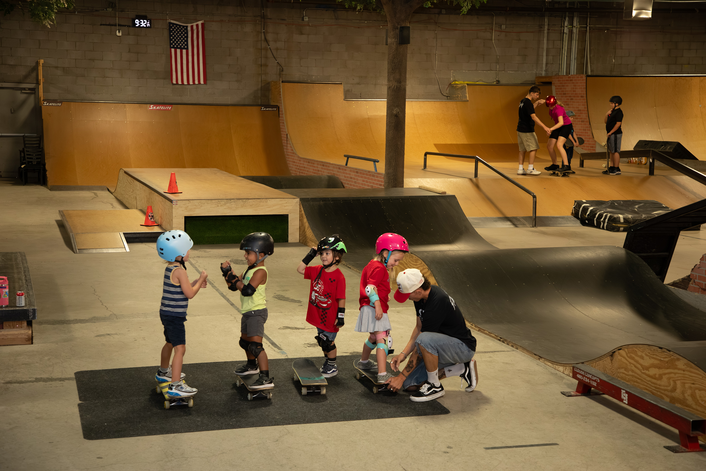 A man helps a young girl onto a skateboard next to three other kids on skateboards, two of whom are playing a game of rock-paper-scissors.