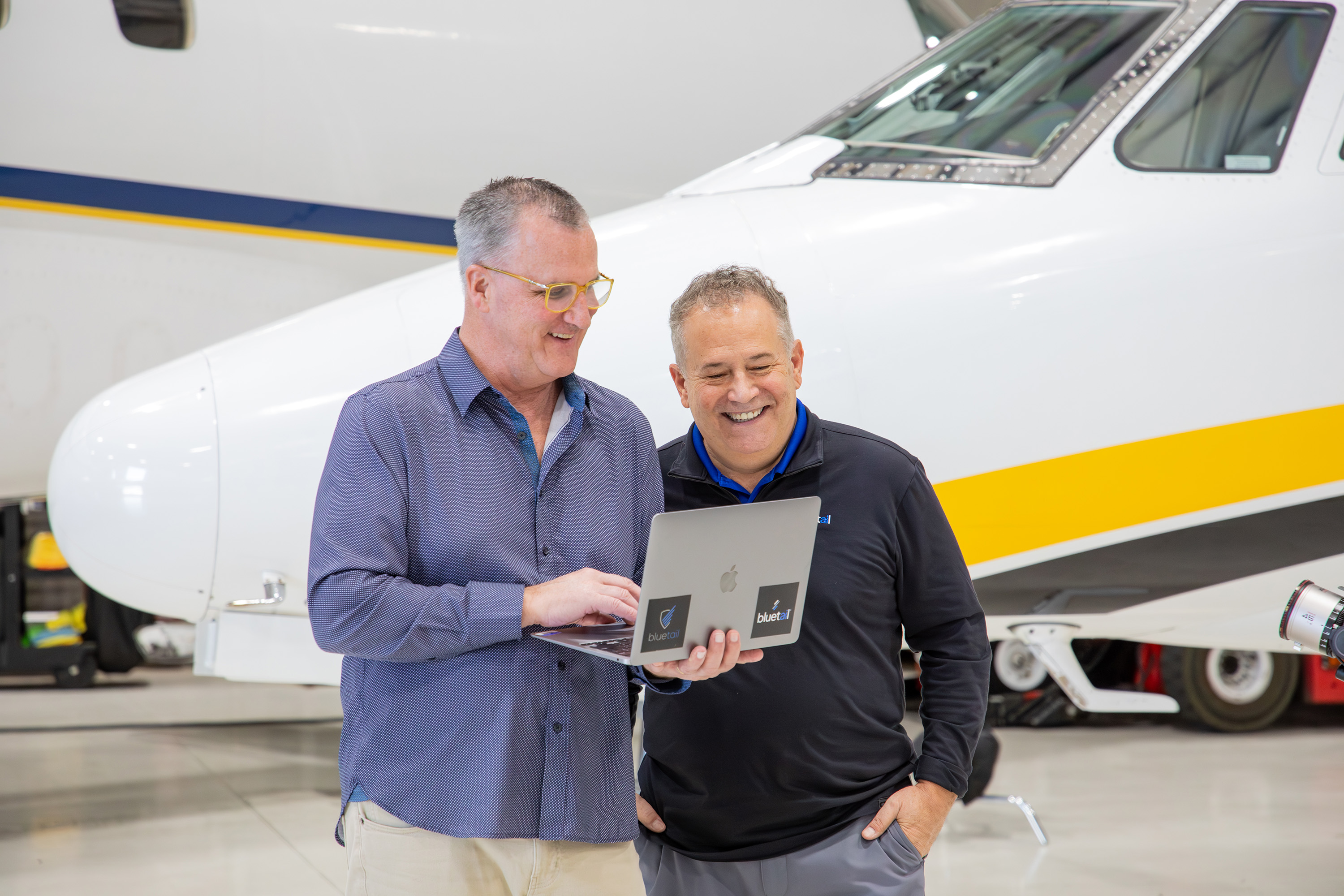 Two men look at a laptop screen while standing in front of the nose of an airplane.