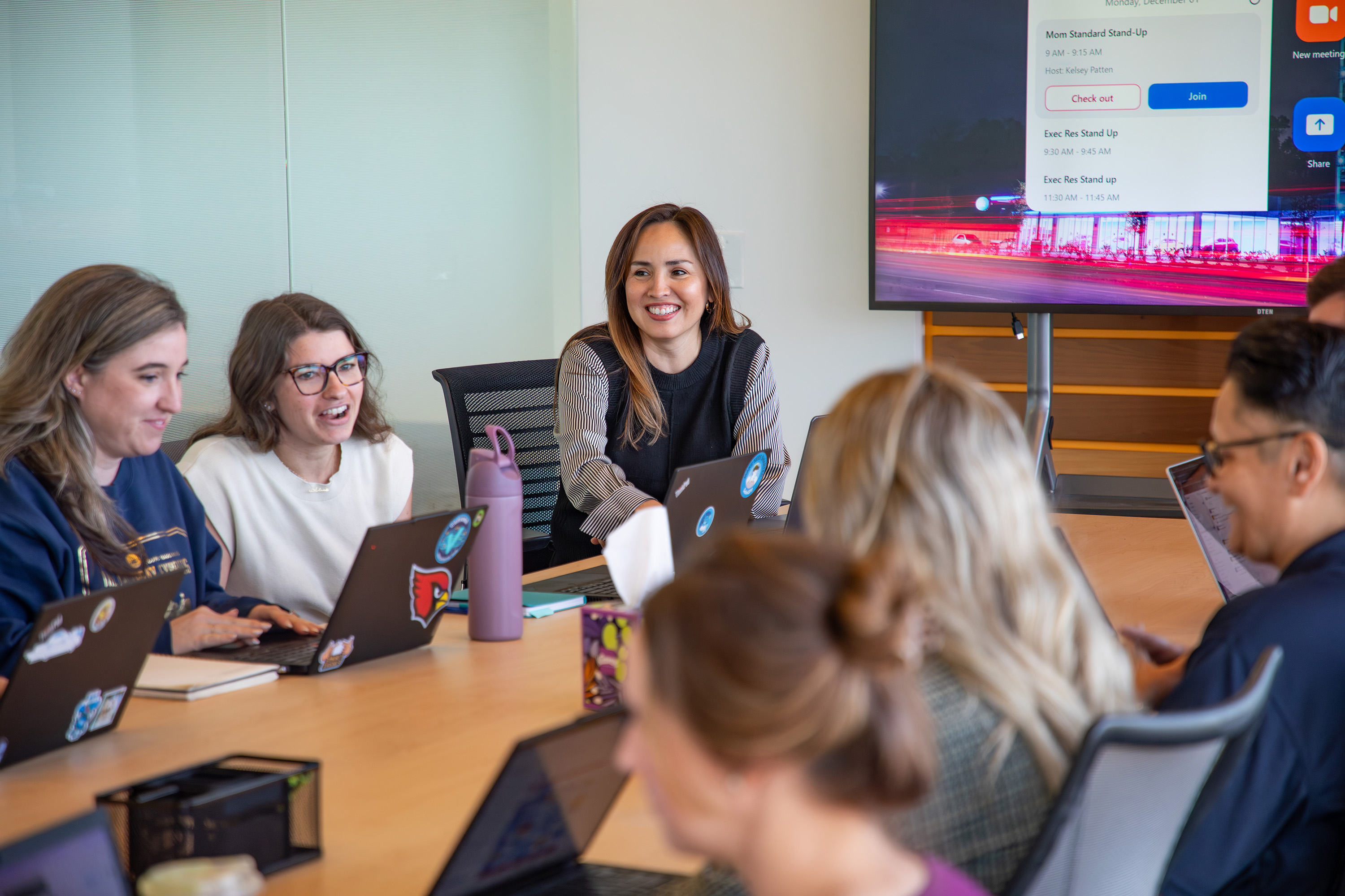 A group of employees sits around a conference table with a TV screen in the background.